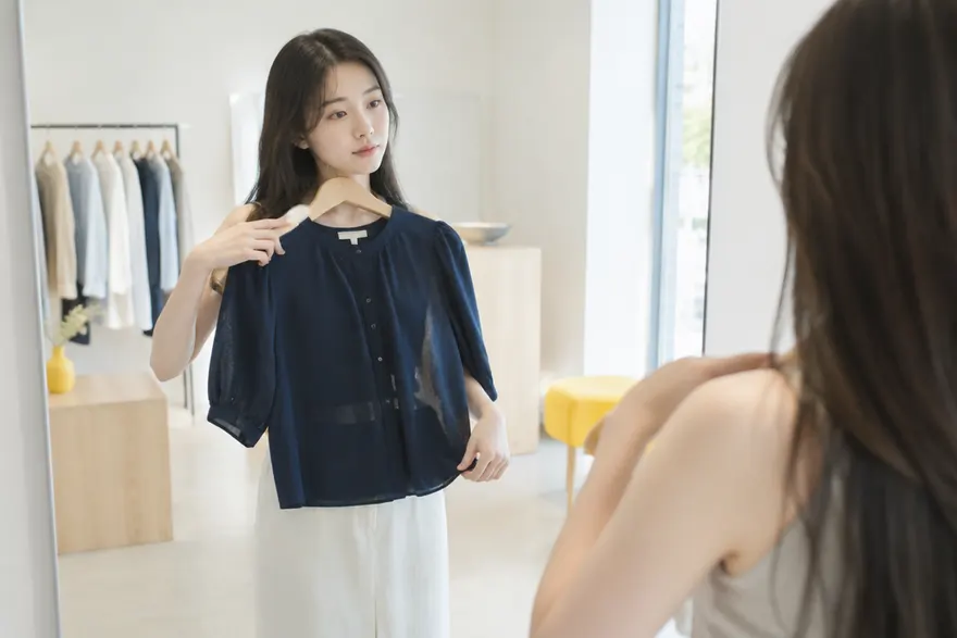 여성이 옷 가게에서 옷걸이에 걸린 원피스를 몸 앞에 대어 보며 크기를 가늠하는 장면